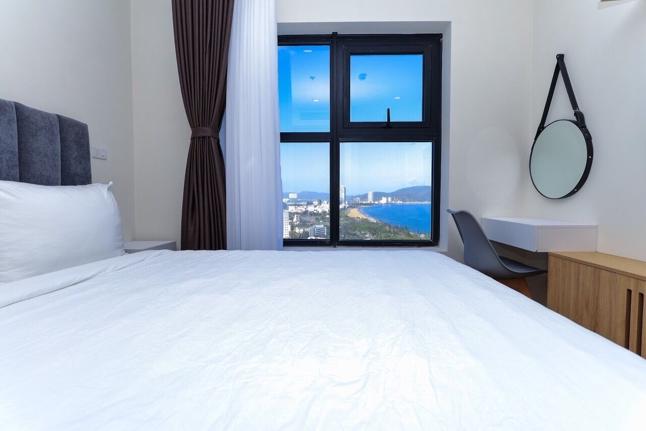 A hotel room with a large bed, a desk, a chair, a round mirror, and a view of the city and sea through a large window.