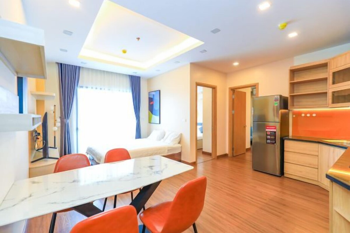 Una habitación de hotel moderna con zona de comedor, sofá, cocina con refrigerador y un amplio ventanal con cortinas.