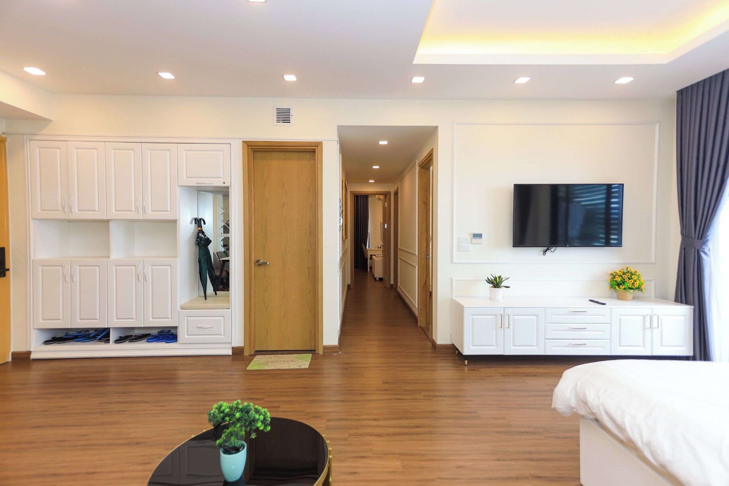 A hotel room with a wooden floor, white furniture, a TV mounted on the wall, and a plant on the table.