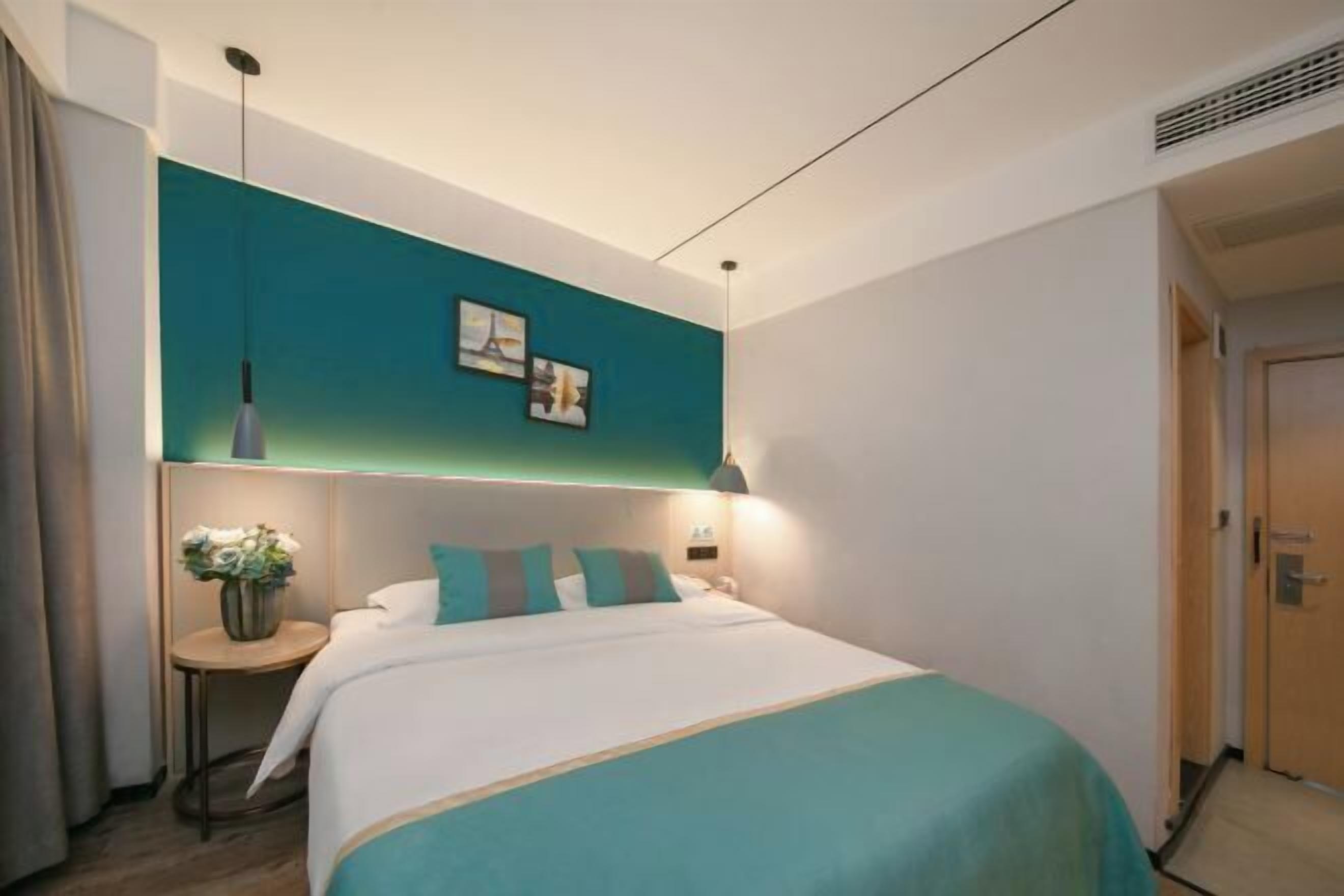 A modern hotel room with a teal accent wall, a bed with white bedding and teal pillows, a bedside table with a vase of flowers, and a picture frame on the wall.