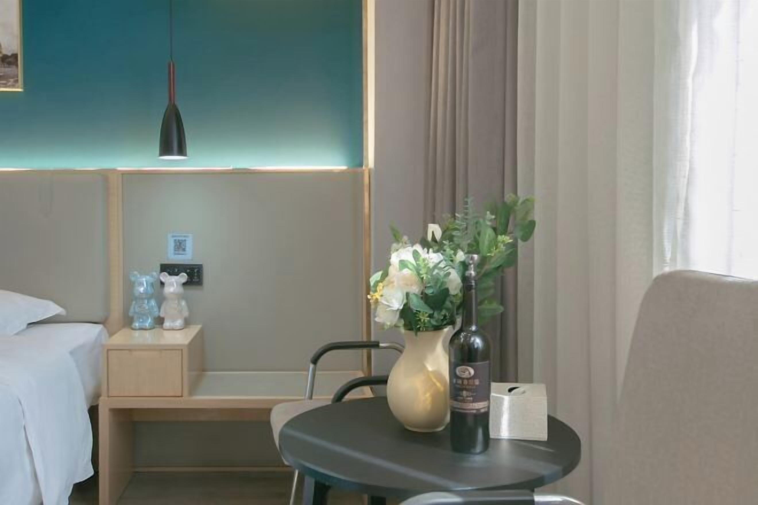 Habitación de hotel con cama, mesita de noche, una pequeña mesa con un jarrón de flores y una botella, una silla y una lámpara fijada a la pared.