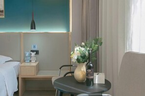 Een hotelkamer met een bed, nachtkastje, een klein tafeltje met een bloevas en een fles, een stoel, en een wandlamp.