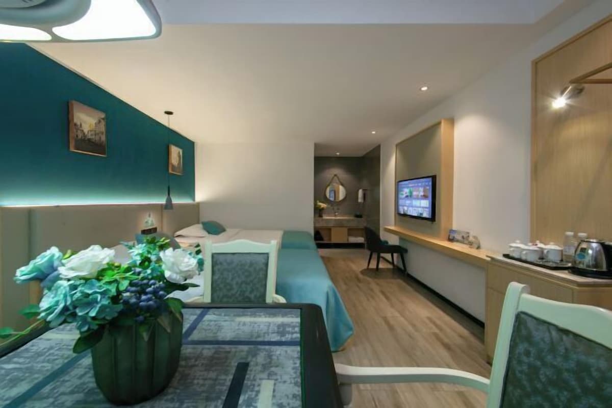 A modern hotel room with a bed, a dining table with a vase of flowers, a TV, and a kitchenette.