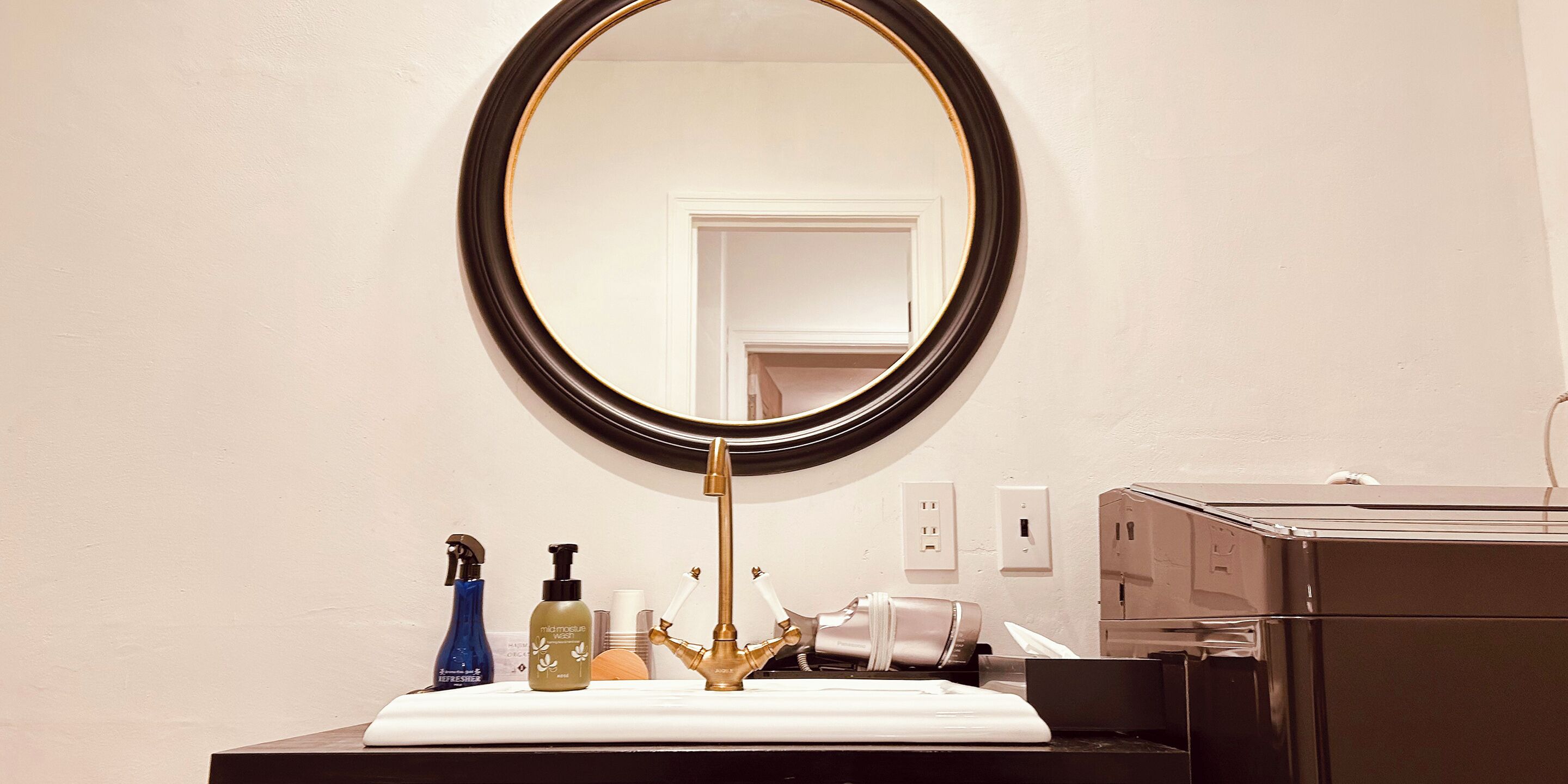 A bathroom with a round mirror, a gold faucet, and toiletries on the counter.