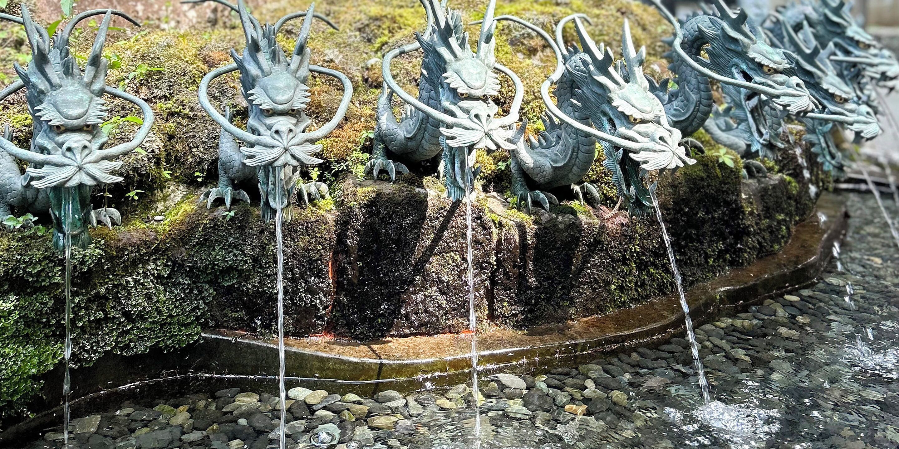 A row of dragon statues with water flowing from their mouths, set against a moss-covered rock surface.