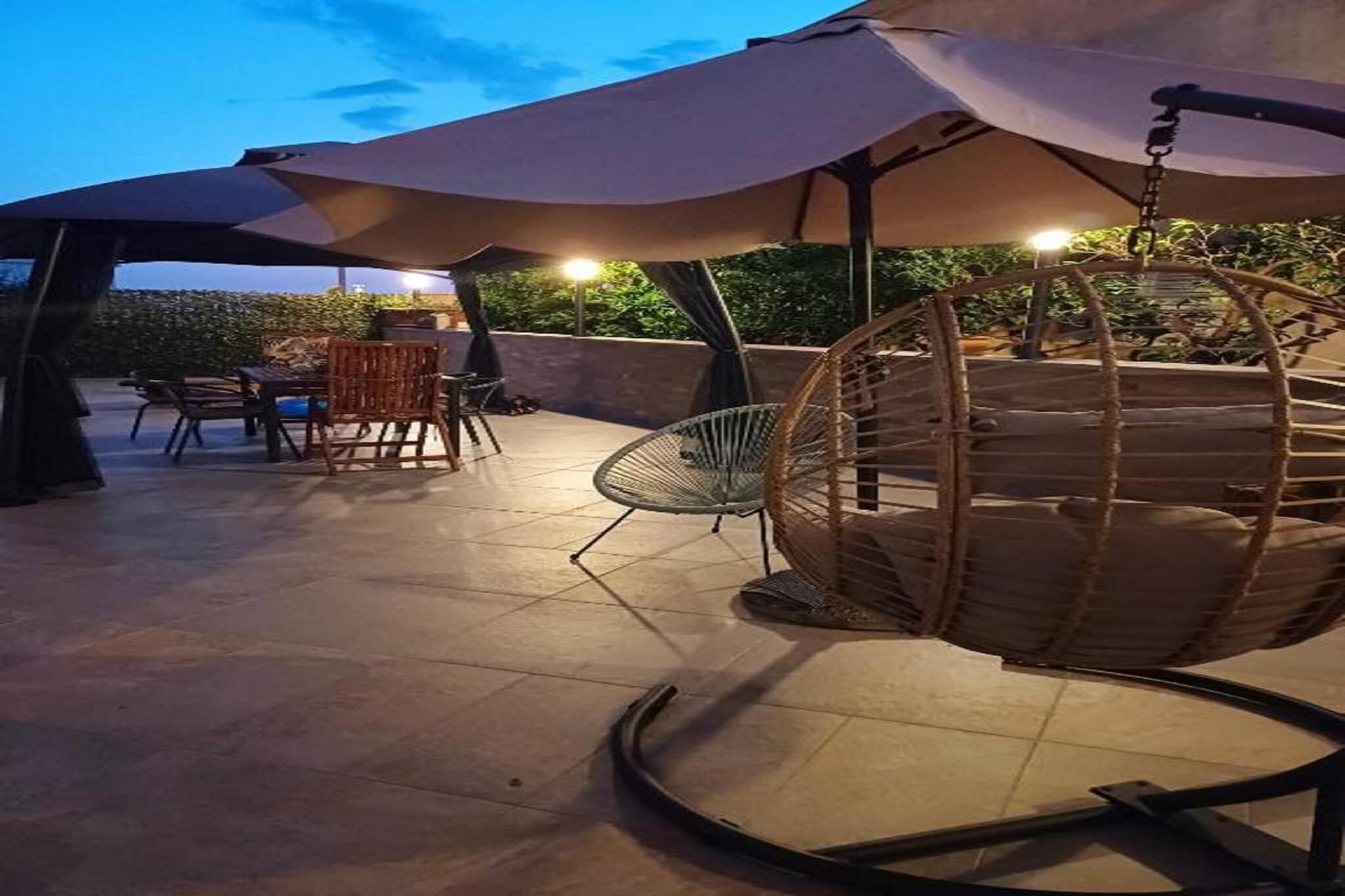 Un área de patio al aire libre con una silla colgante, una mesa con sillas y un amplio paraguas que da sombra.