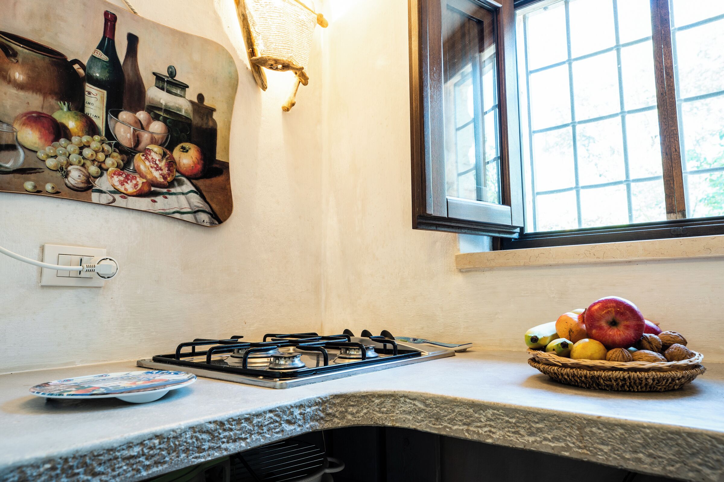 Una cocina con estufa a gas, frutero, un cuadro en la pared y ventana con vistas.