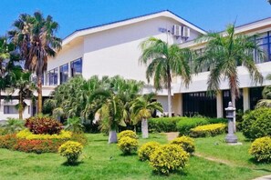 Ein Hotel mit heller Fassade, mehreren Stockwerken und einem gepflegten Garten mit Palmen und Sträuchern.