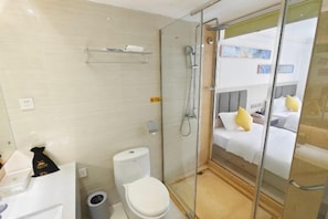 A compact hotel room with a glass shower enclosure, a white toilet, and a bed with yellow pillows.