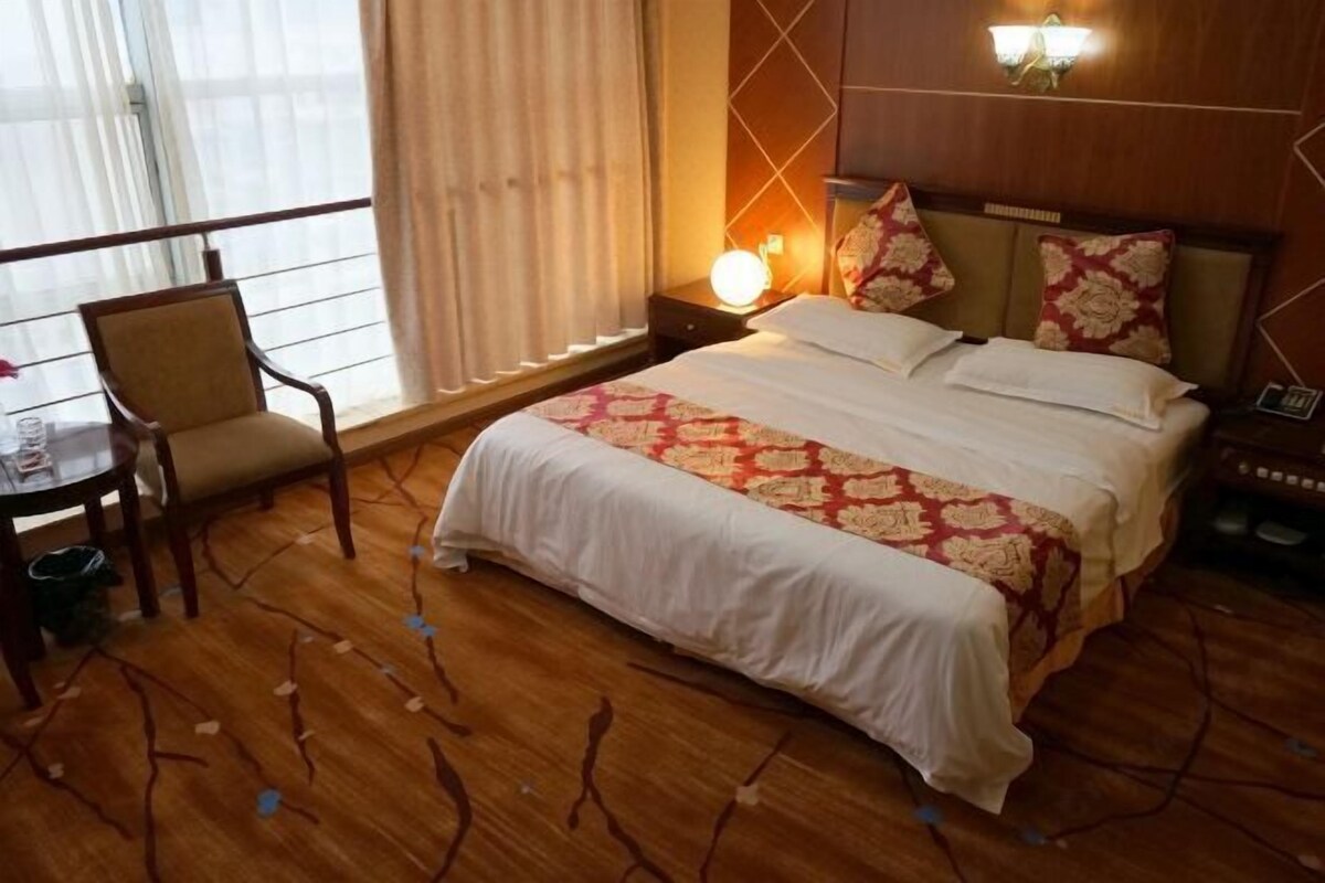 A hotel room with a large bed, a bedside table with a lamp, a chair, and a window with curtains.