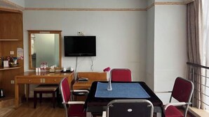 Una habitación con escritorio de madera, televisor montado en la pared, mesa de comedor con sillas y una ventana con cortinas.