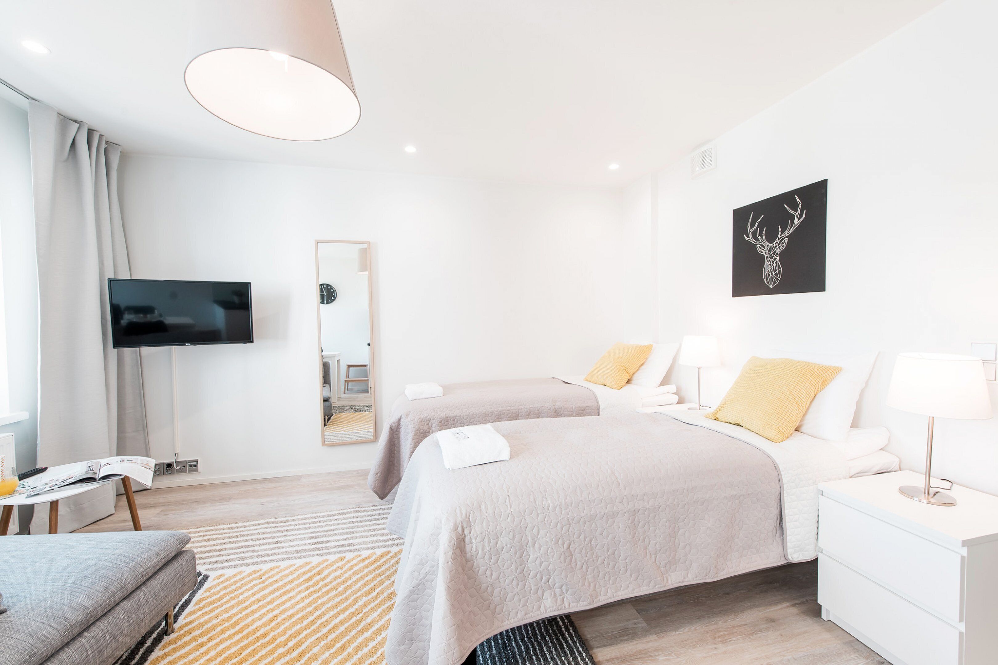 A modern hotel room with two beds, a flat-screen TV, a mirror, and a wall-mounted deer head artwork.