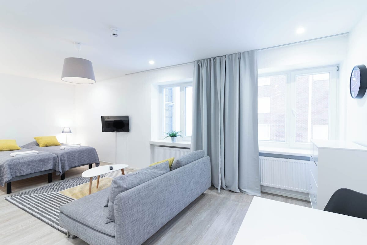Une chambre d’hôtel moderne avec deux lits simples, un canapé gris, une télévision et de grandes fenêtres équipées de rideaux.