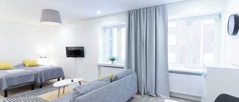 A modern hotel room with two single beds, a grey sofa, a TV, and large windows with curtains.