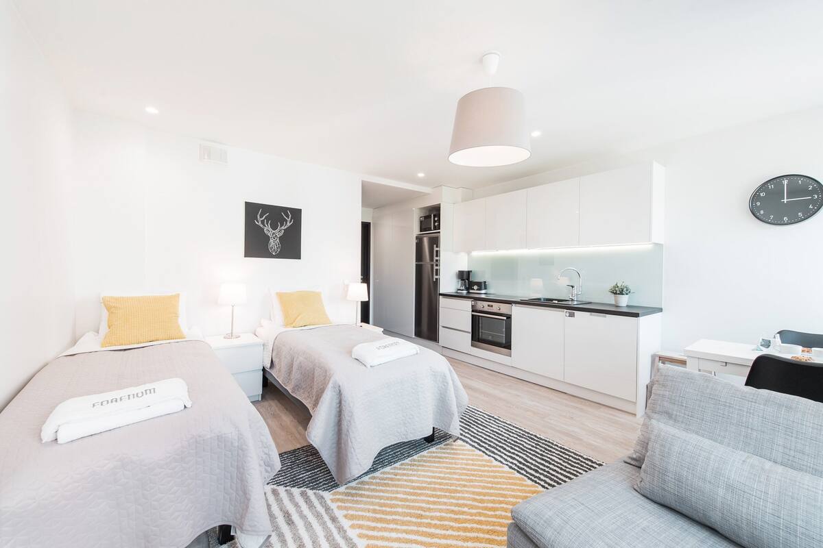 Une chambre d’hôtel moderne avec deux lits, un coin cuisine, un espace repas et une grande horloge au mur.