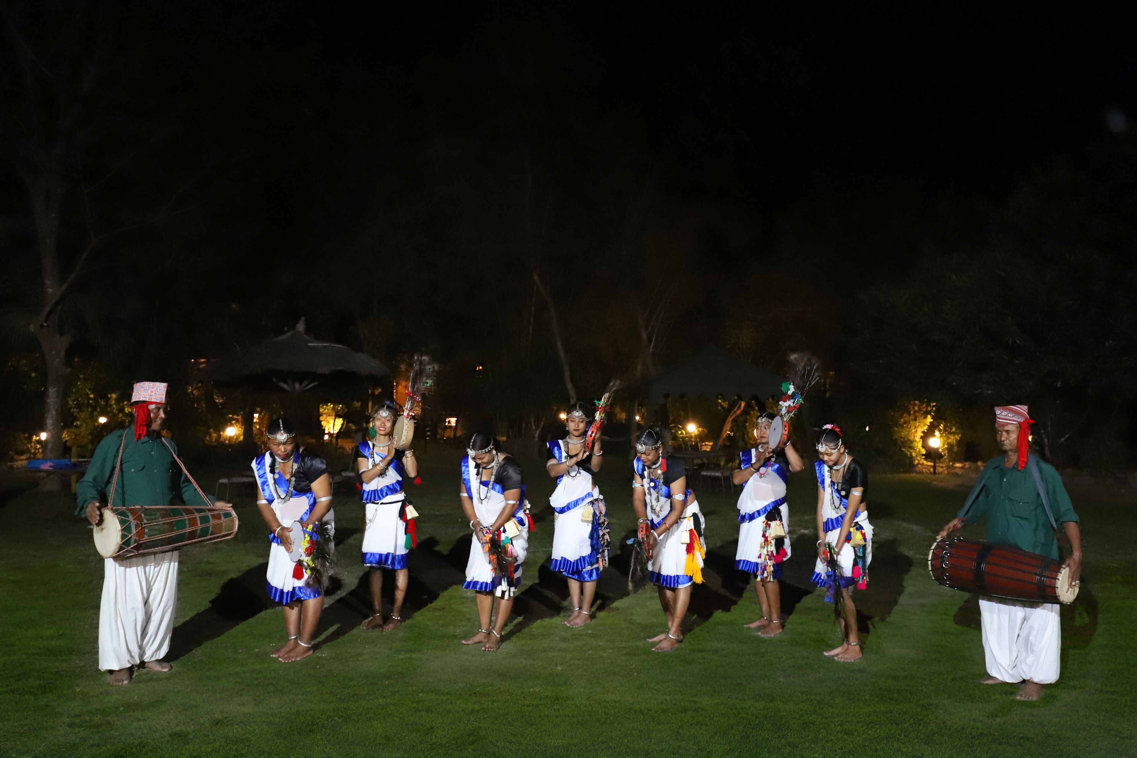 Un grupo de personas vestidas con trajes tradicionales actuando en un escenario al aire libre sobre césped, de noche.