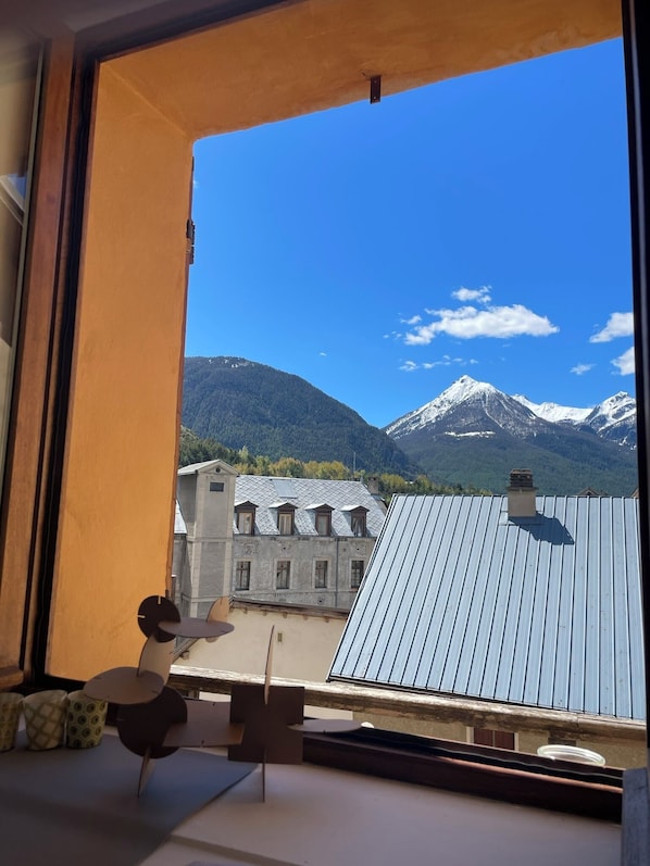 A view from a window with a metal roof, overlooking a building with multiple windows and a mountain range in the background.