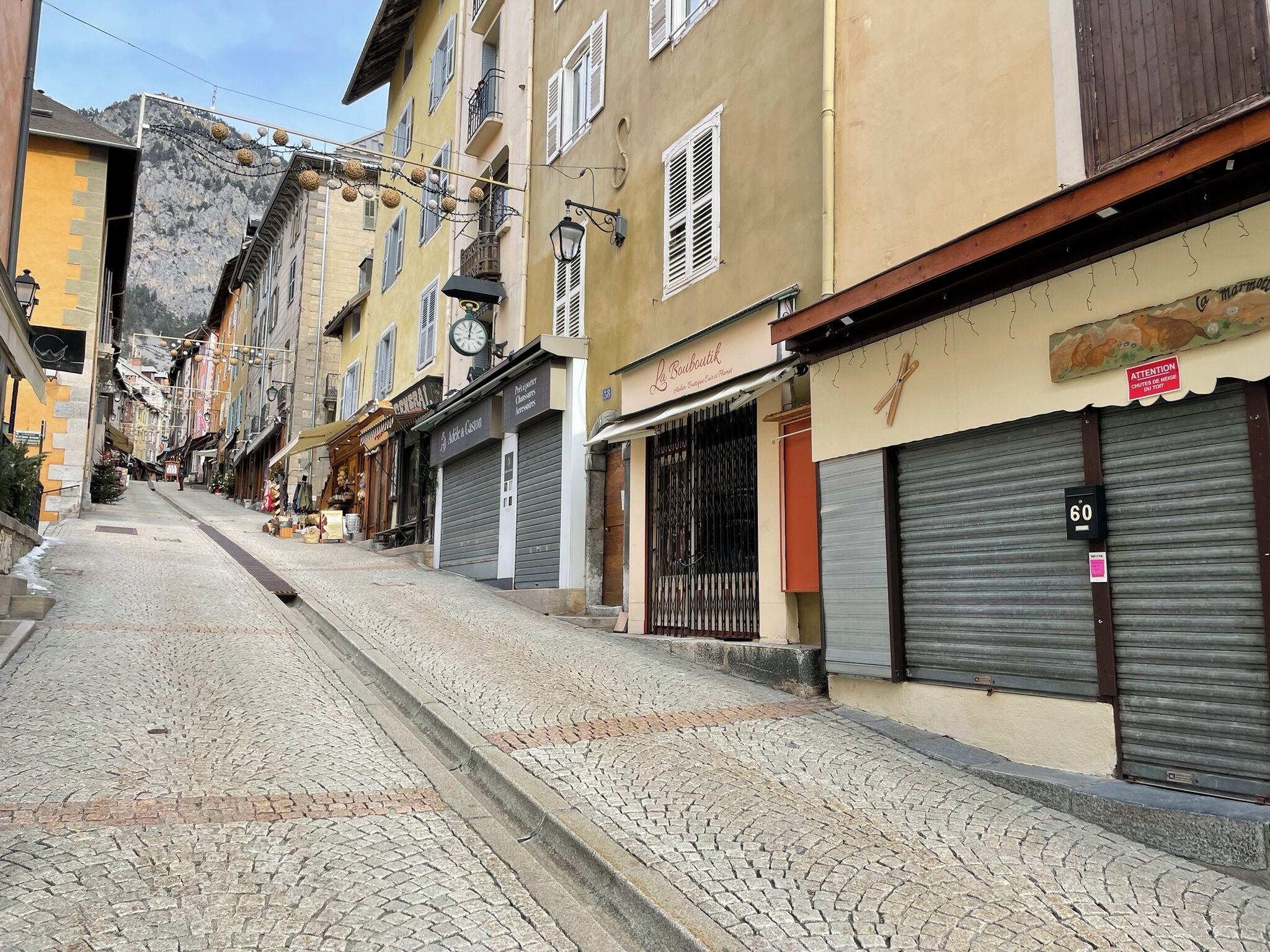 A cobblestone street lined with buildings, some with closed shutters, indicating shops or businesses.