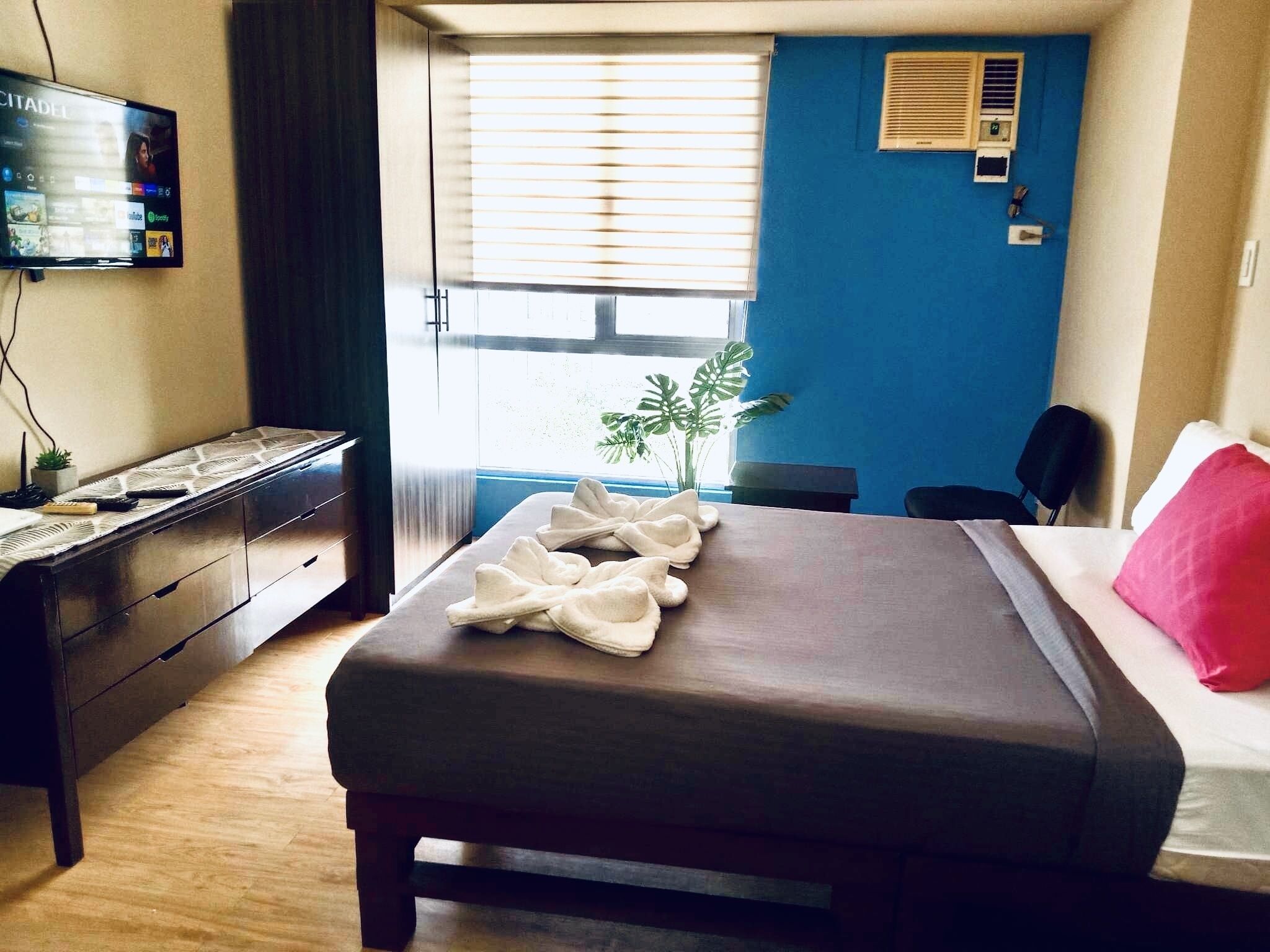 Ein Hotelzimmer mit einem Bett, einem Schreibtisch mit Computer, einem Fernseher und einem Fenster mit Jalousien.