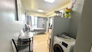 A compact room with a bed, a small table with chairs, a washing machine, and a window with curtains. - IT Park Studio w/ washing machine (Cebu City)