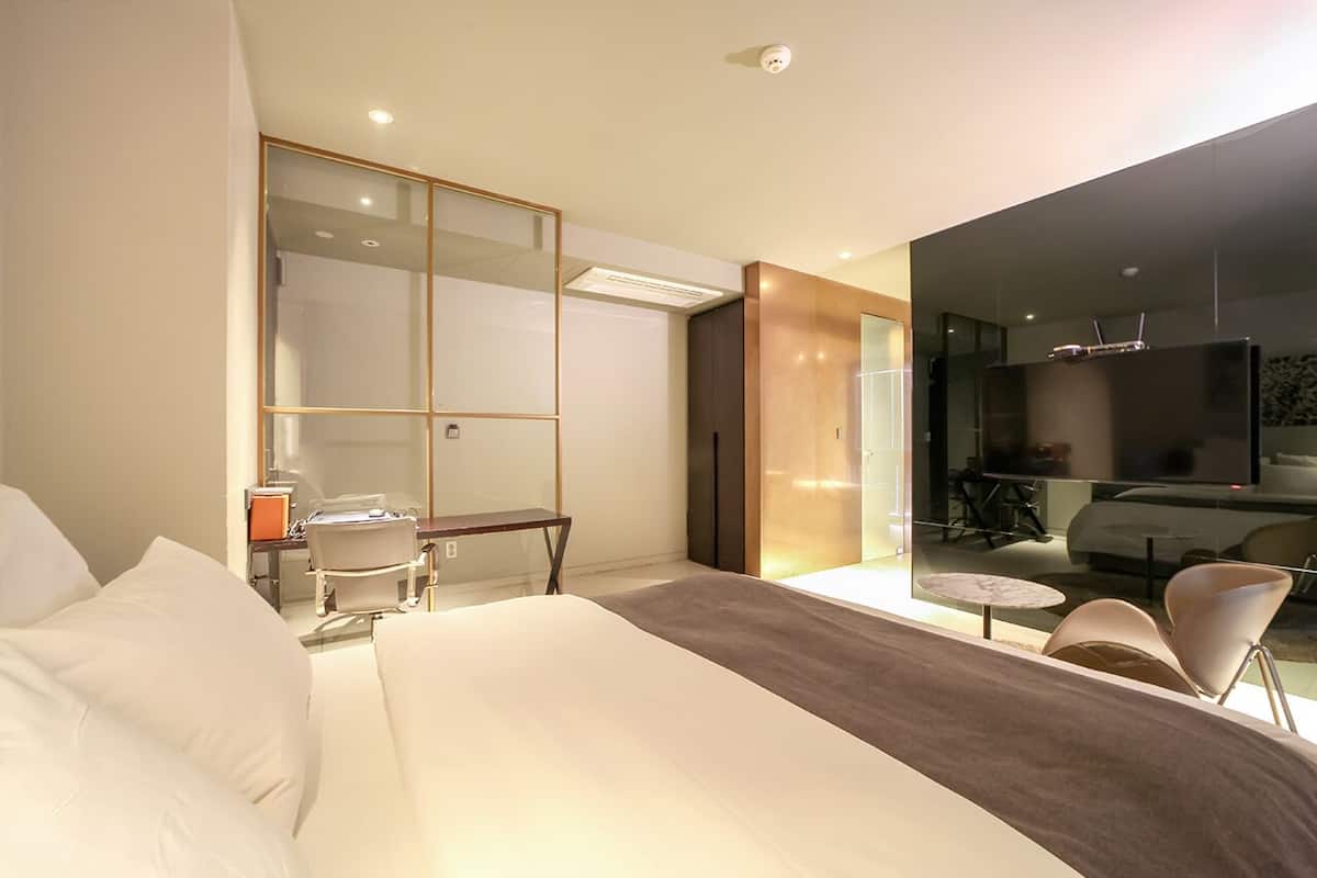 A modern hotel room with a large bed, a desk, a chair, a TV, and a glass partition.