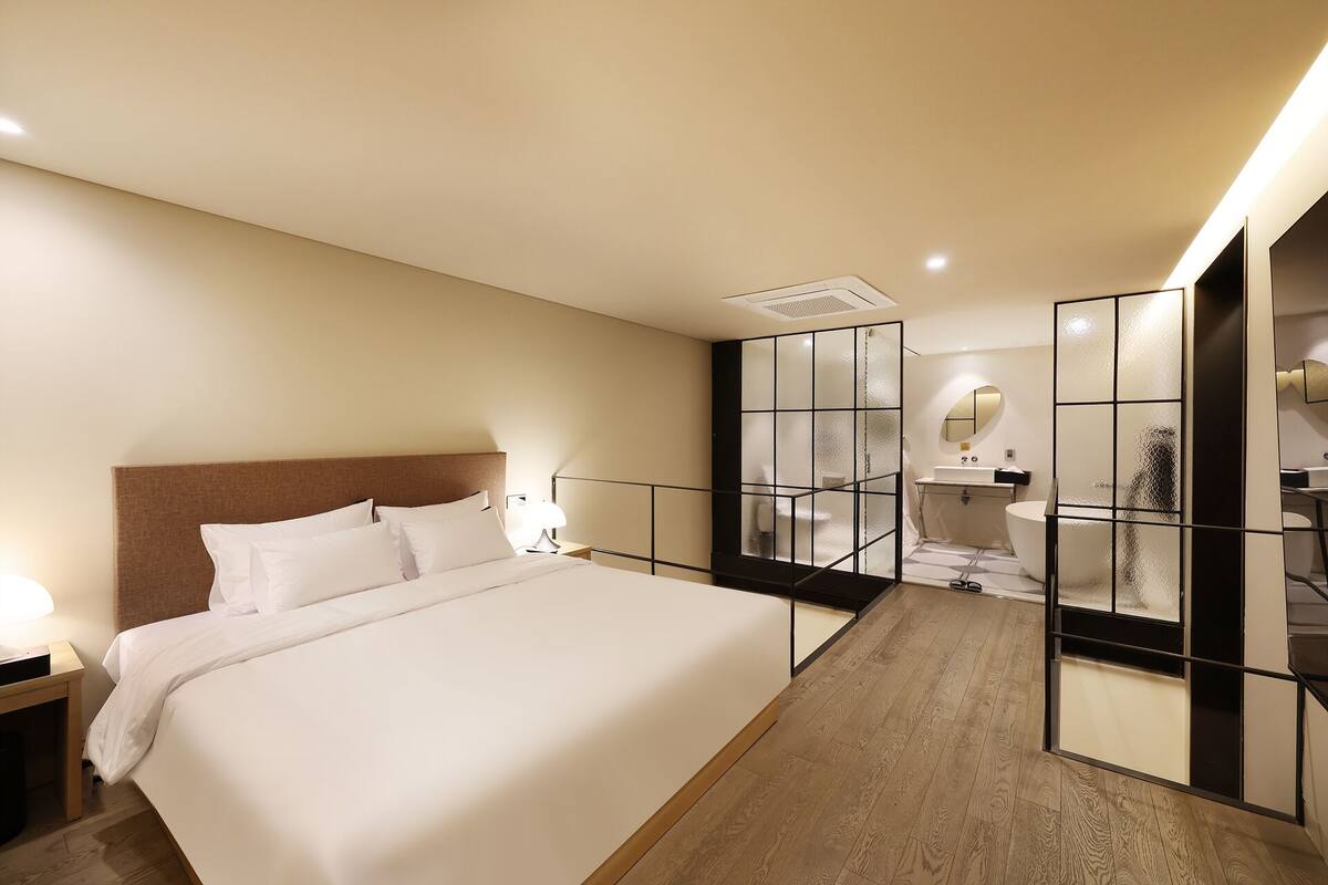 A hotel room with a large bed, a nightstand, a bathroom with a mirror and sink, and a wooden floor.
