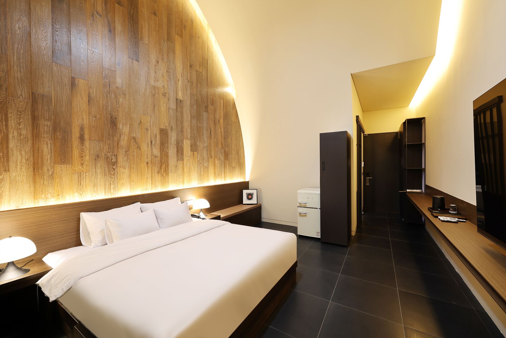 Ein Hotelzimmer mit einem großen Bett, Nachttischen mit Lampen, einer Wand mit Holzakzenten, einem Kühlschrank, einem Schreibtisch mit Stuhl und einem Spiegel.