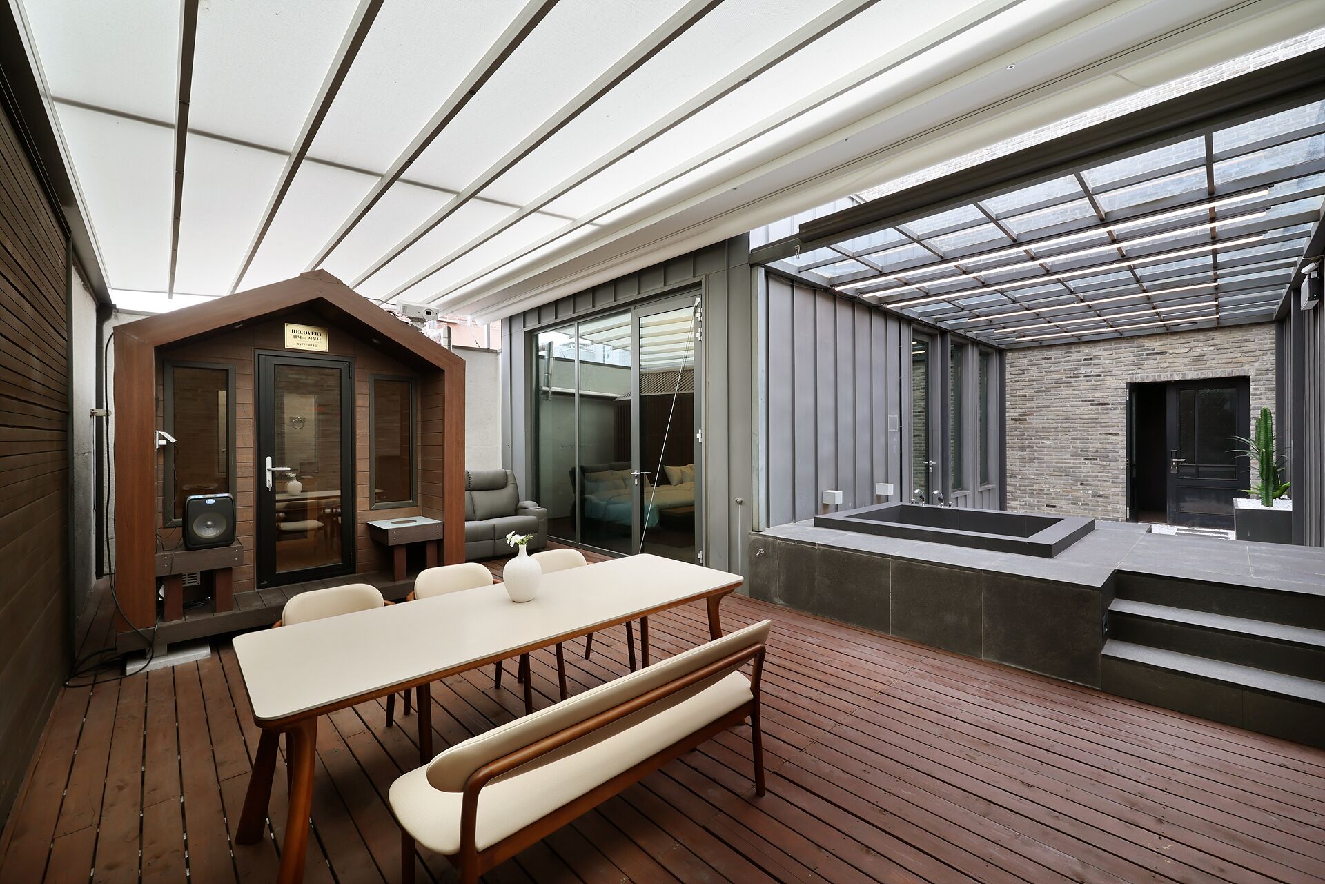 Un área de comedor al aire libre moderna, con una mesa y bancos de madera, una cocina con paredes de vidrio y una pared de ladrillo.