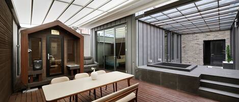 Un área de comedor al aire libre moderna, con una mesa y bancos de madera, una cocina con paredes de vidrio y una pared de ladrillo.