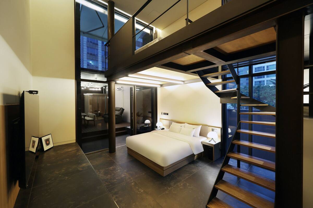 A modern bedroom with a bed, a desk, and a chair. There is a staircase leading to an upper level.