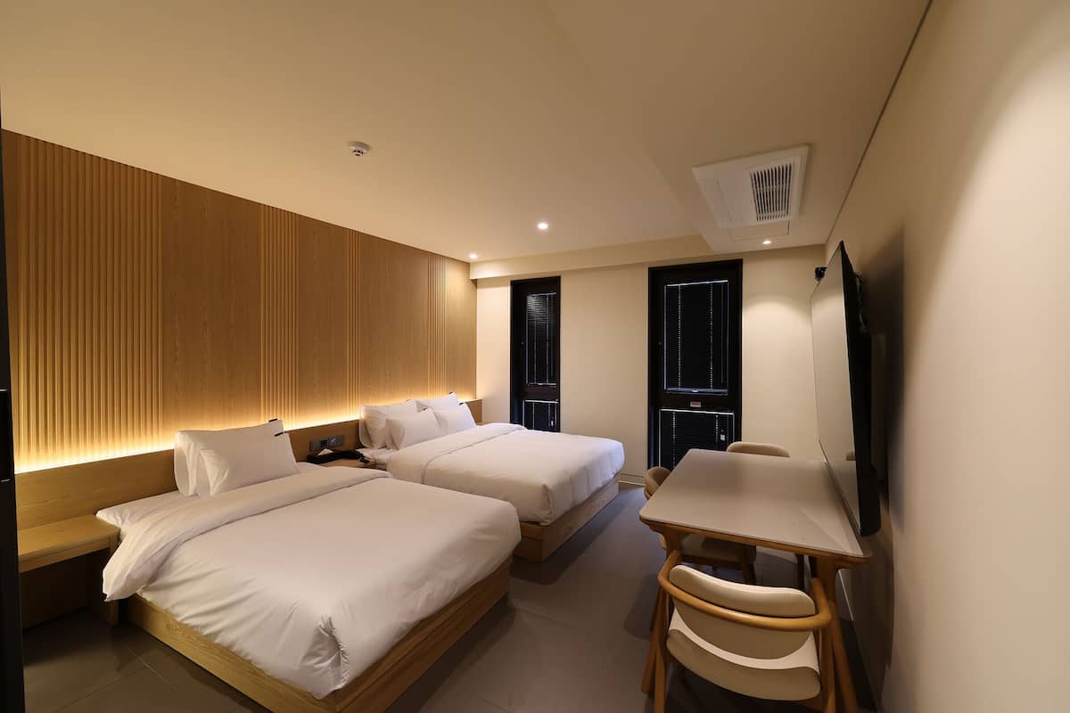 A hotel room with two beds, a desk, a chair, a TV, and a ceiling-mounted air conditioning unit.