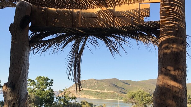 A rustic outdoor seating area with a thatched roof, surrounded by natural landscape and a view of hills and water.