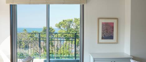 A room with a large sliding glass door leading to a balcony with a view of the sea and trees. There is a framed picture on the wall and a small white bedside table with a drawer.