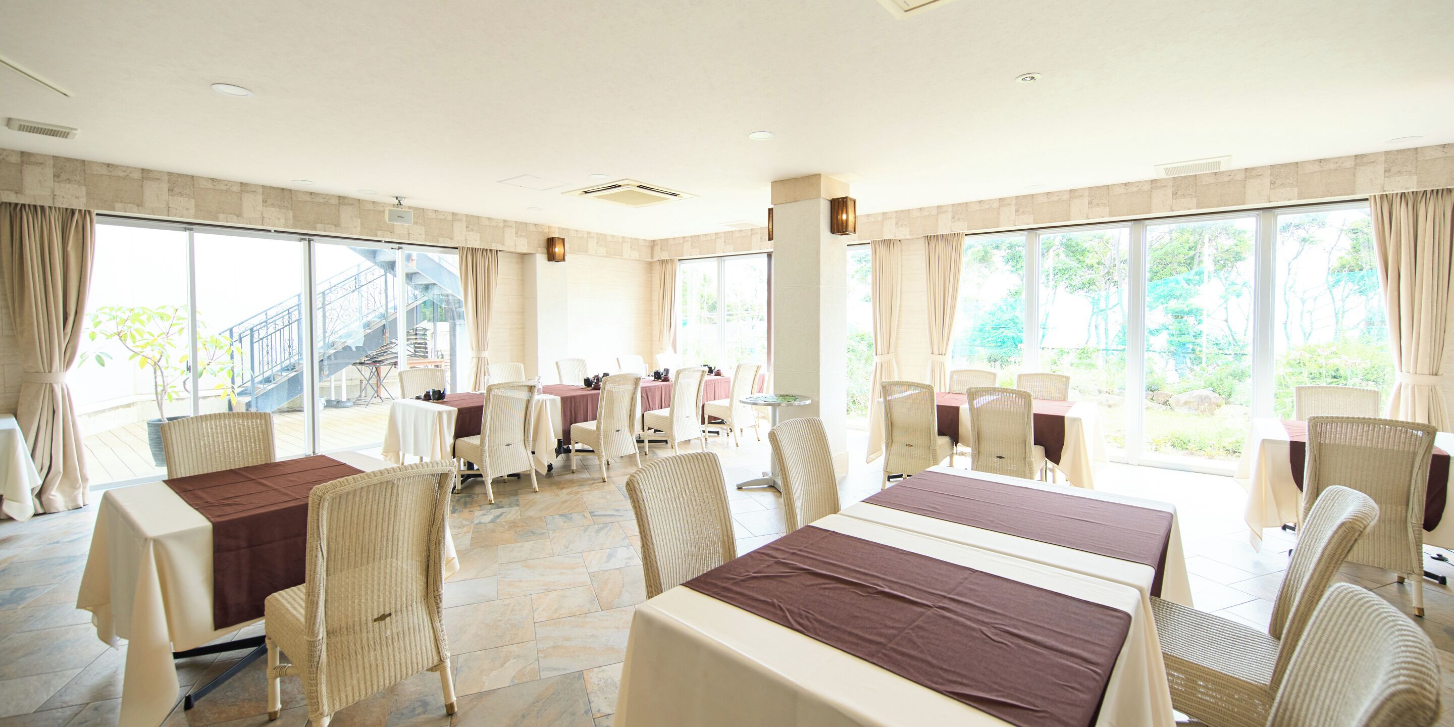 A spacious conference room with multiple tables and chairs, large windows with curtains, and a stone-tiled floor.