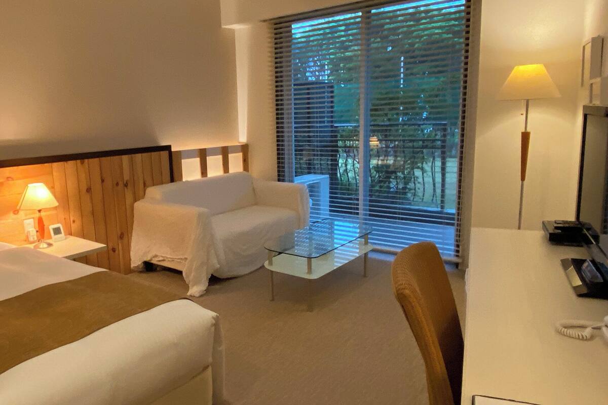 A hotel room with a bed, a chair, a small table, a desk, a lamp, and a window with blinds.