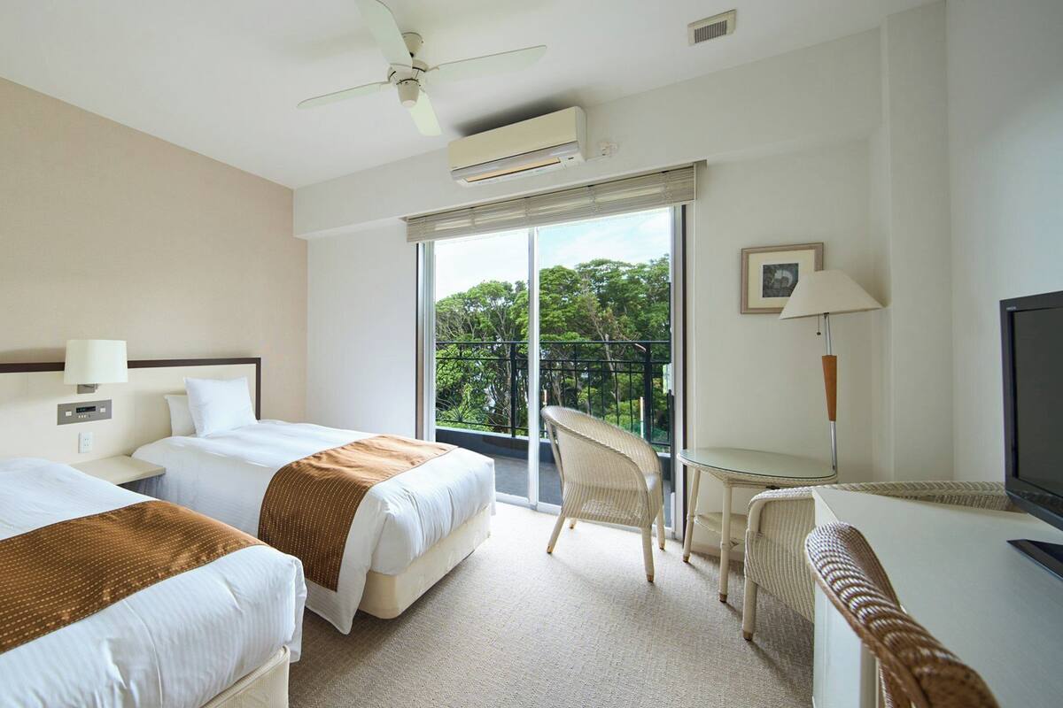 A hotel room with two beds, a ceiling fan, an air conditioning unit, a balcony with a chair, and a television.