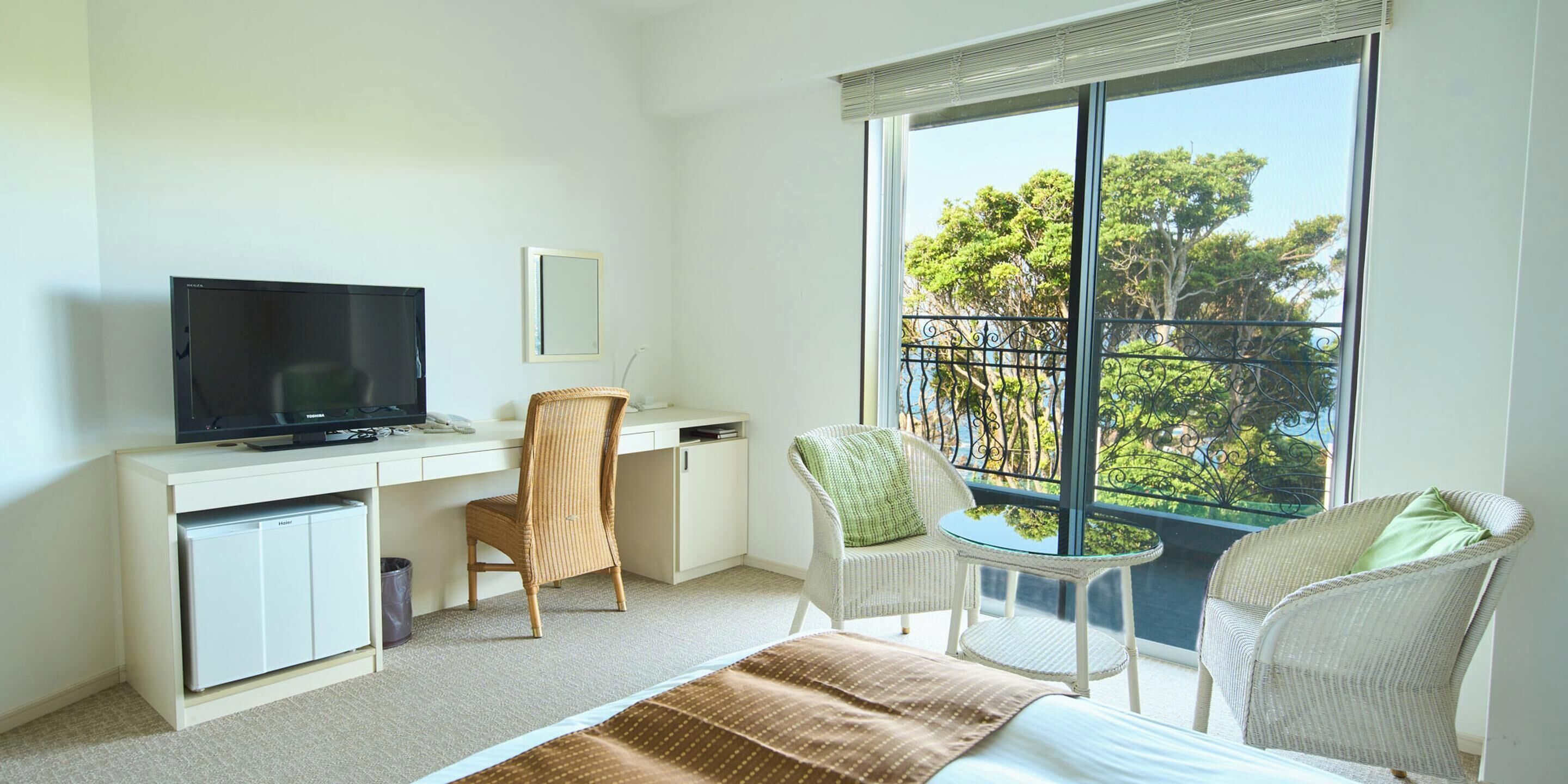 Ein Hotelzimmer mit Bett, Schreibtisch mit Fernseher, Stuhl, kleinem Tisch und Fenster mit Blick auf Bäume.
