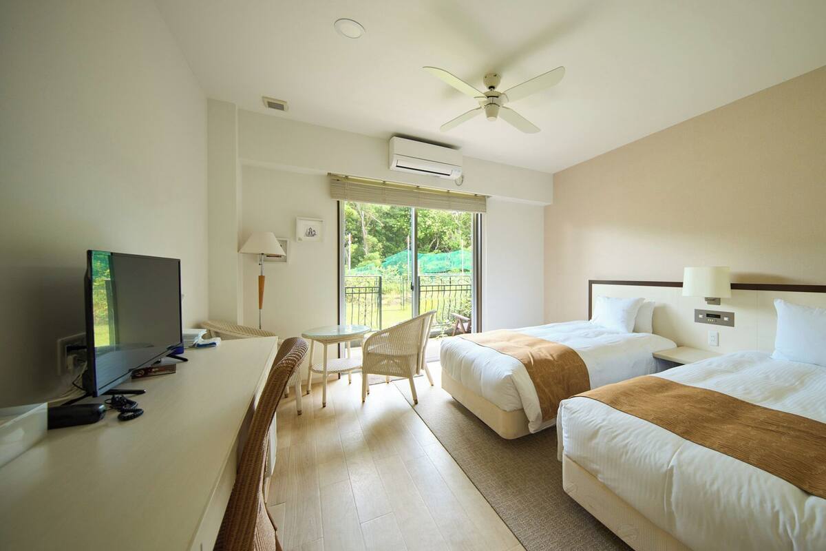 A hotel room with two beds, a desk with a TV, a chair, a ceiling fan, and a view of the outdoors.