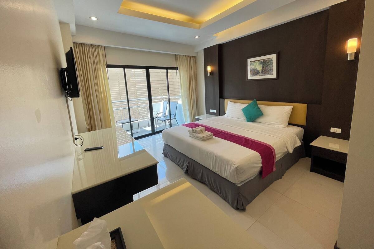 A modern hotel room with a large bed, a desk, and a balcony with a view.