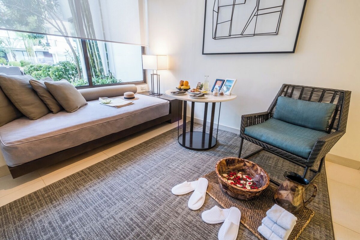 A modern room with a sofa, a wicker chair, a small table with food and drinks, and a window with a view of greenery.