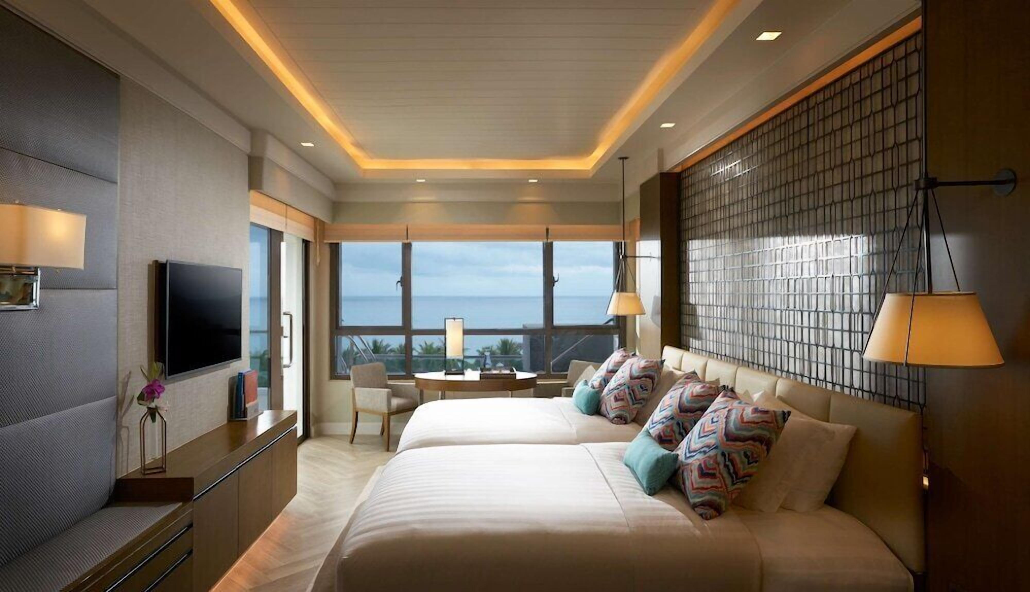 A modern hotel room with a large bed, a TV, a desk, and a view of the ocean.