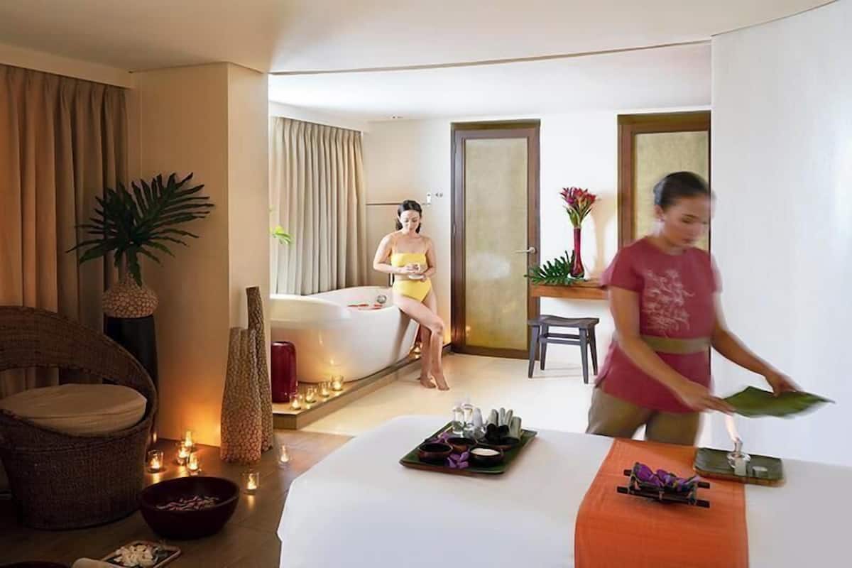 A hotel room with a bed, a sitting area with a chair, a dining table with a tray of food, and a bathroom with a bathtub.