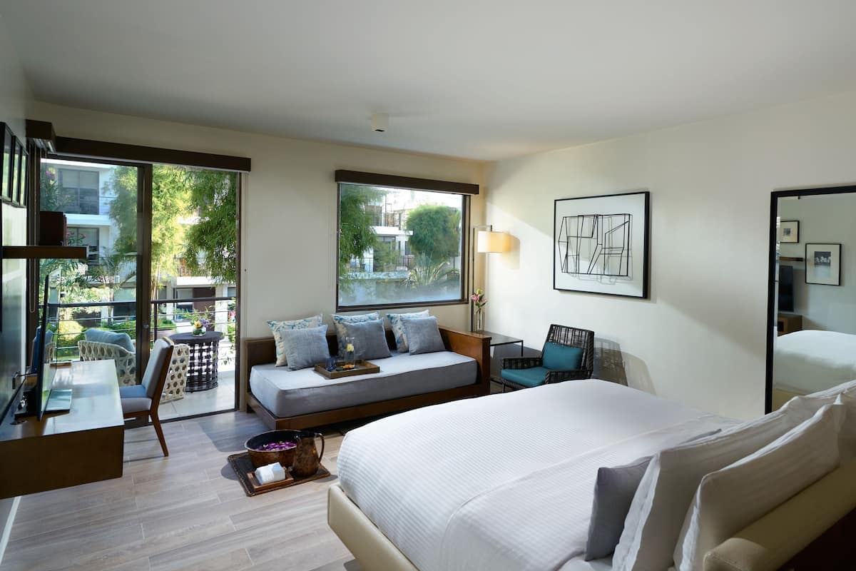 A hotel room with a large bed, a sofa, a chair, a desk, and a large window offering a view of greenery.