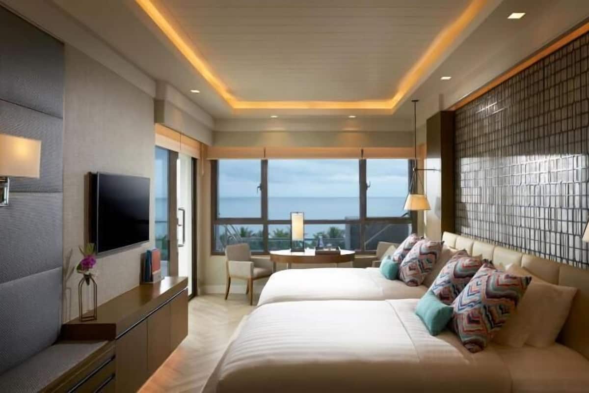 A hotel room with two beds, a TV, a desk, and a view of the ocean.