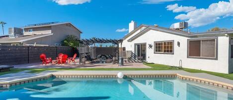 A swimming pool with a patio area, a covered seating area, and a white building with a garage.