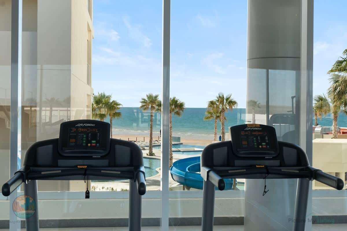 Dos bicicletas estáticas con pantallas digitales, situadas frente a un amplio ventanal con vista a una playa y palmeras.