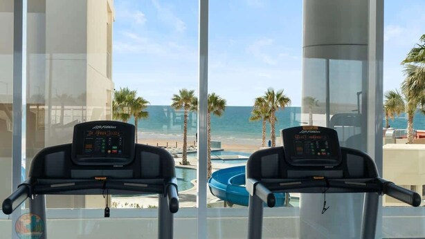 Two exercise bikes with digital displays, positioned in front of a large window overlooking a beach and palm trees. - Encantame Towers | Ground Floor| Lazy River| Pool (Playa Encanto)
