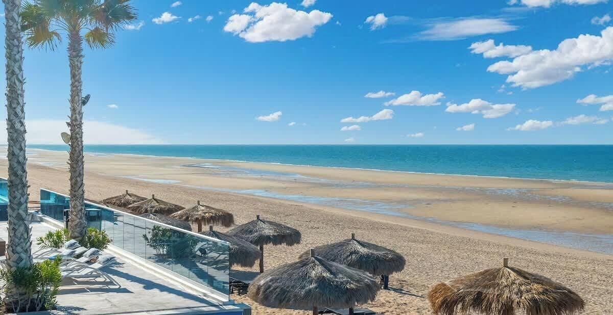 Uitzicht op een strand met palmbomen, een zwembad en rieten parasols.