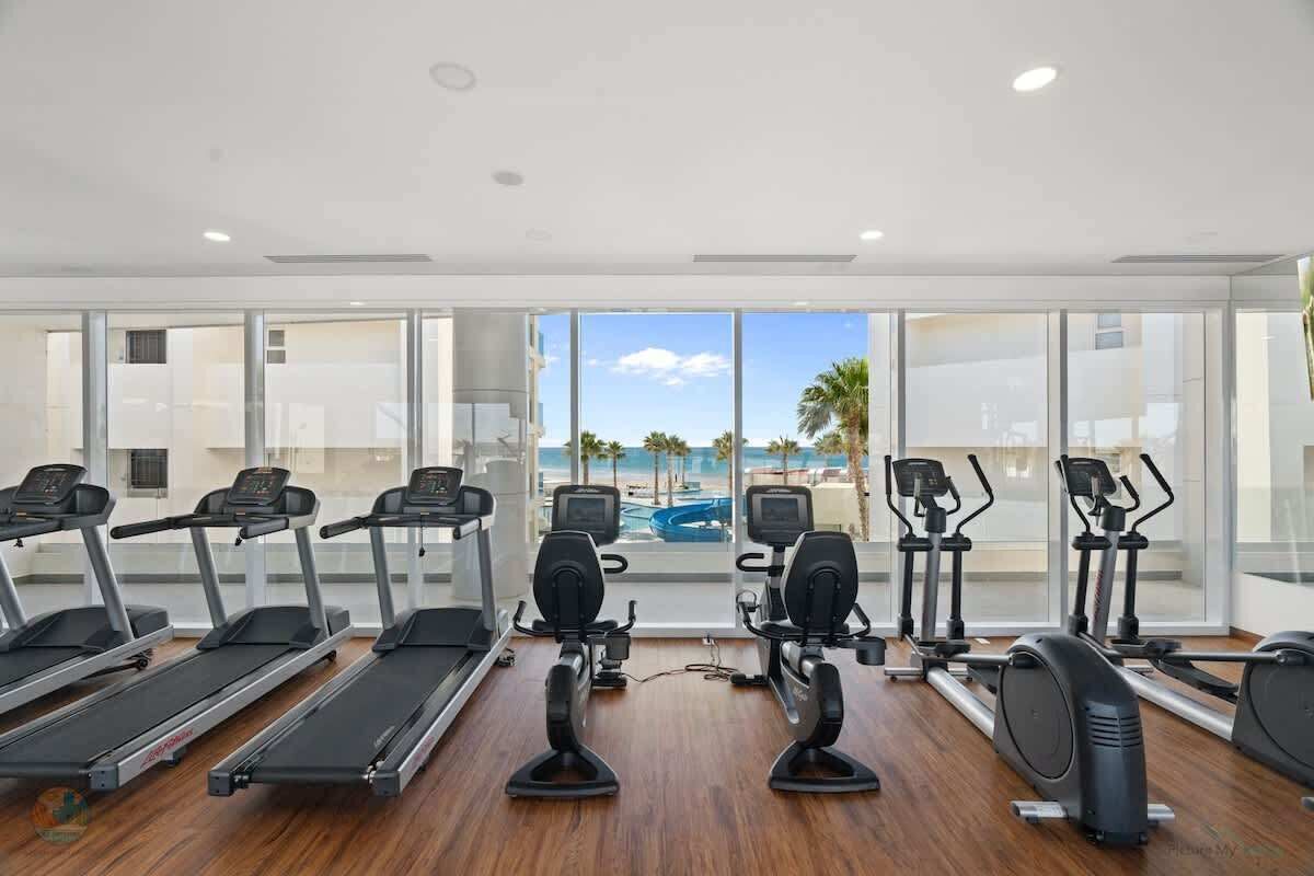 A gym with treadmills, elliptical machines, and stationary bikes, overlooking a pool and palm trees.