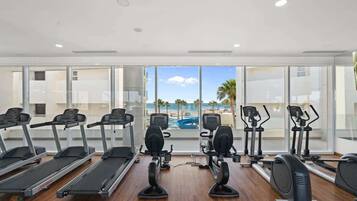 A gym with treadmills, elliptical machines, and stationary bikes, overlooking a pool and palm trees.